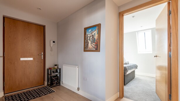 Un couloir avec une porte en bois, un radiateur, un tableau et, au fond, on aperçoit une chambre avec un lit et une fenêtre.