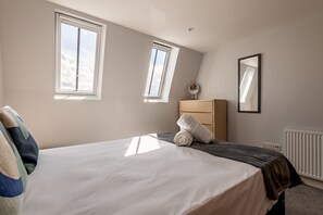 A bedroom with a bed, two windows, a dresser, a mirror, and a radiator. - Century Court (No 10) | St Andrews (St Andrews)