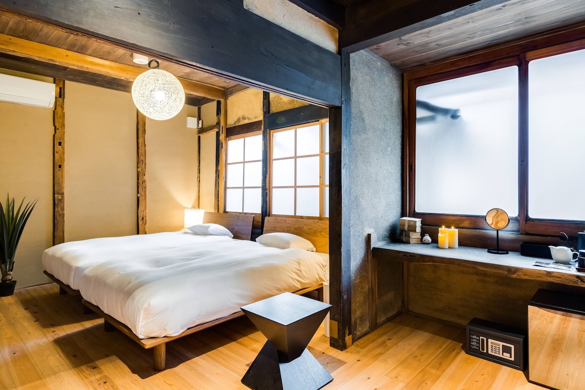 Un dormitorio con cama, mesita de noche, ventana y una pequeña zona de escritorio con lámpara y velas.