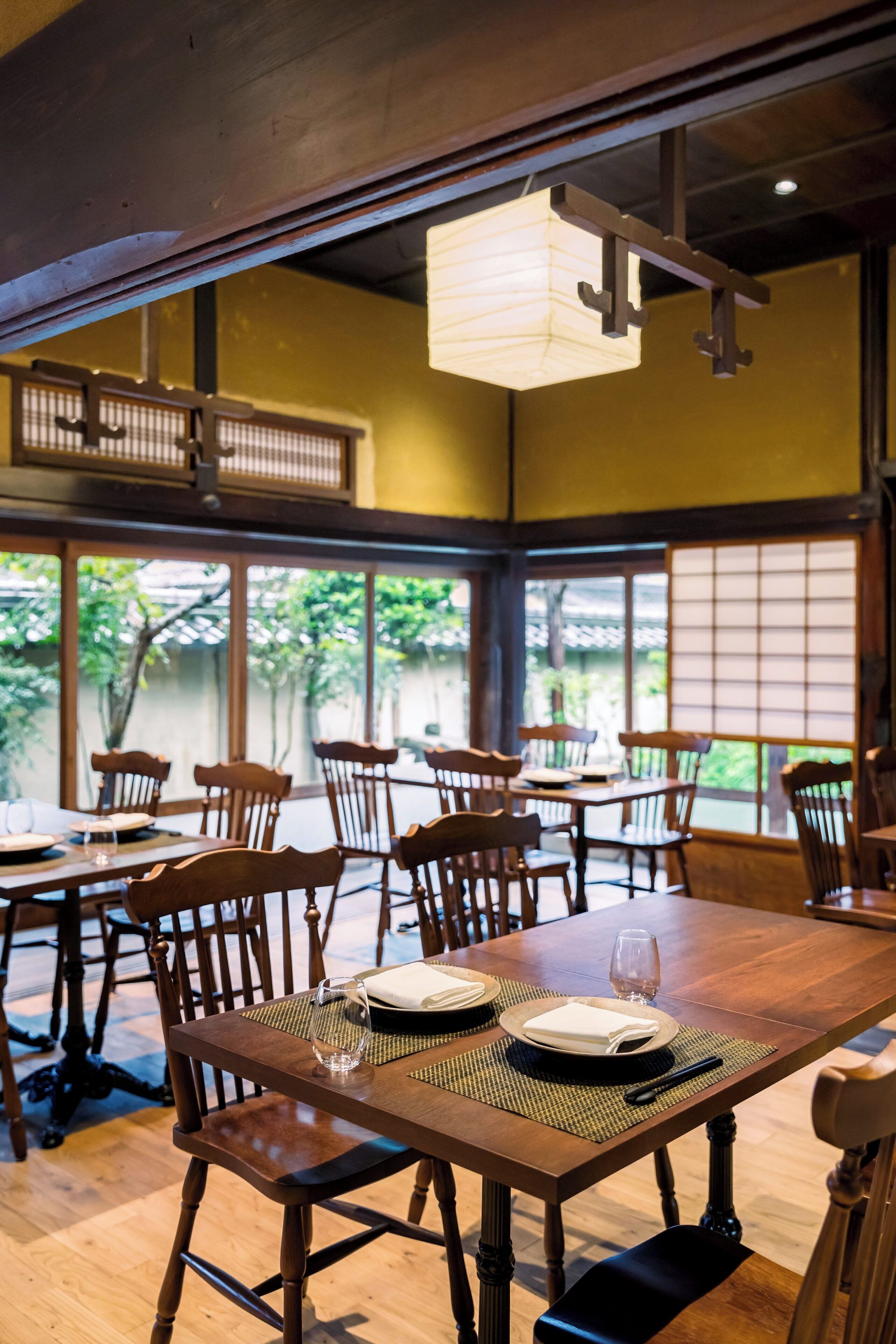A restaurant interior with wooden tables and chairs, a hanging light fixture, and large windows with a view of greenery outside.