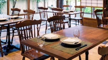 A restaurant interior with wooden tables and chairs, a hanging light fixture, and large windows with a view of greenery outside.