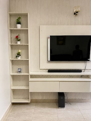 A modern living room with a flat-screen TV mounted on a white wall, a built-in shelf unit with decorative items, and a black speaker on the floor. - Grand 4BR Luxury Duplex near Airport – 15 mins from airport (Benin)