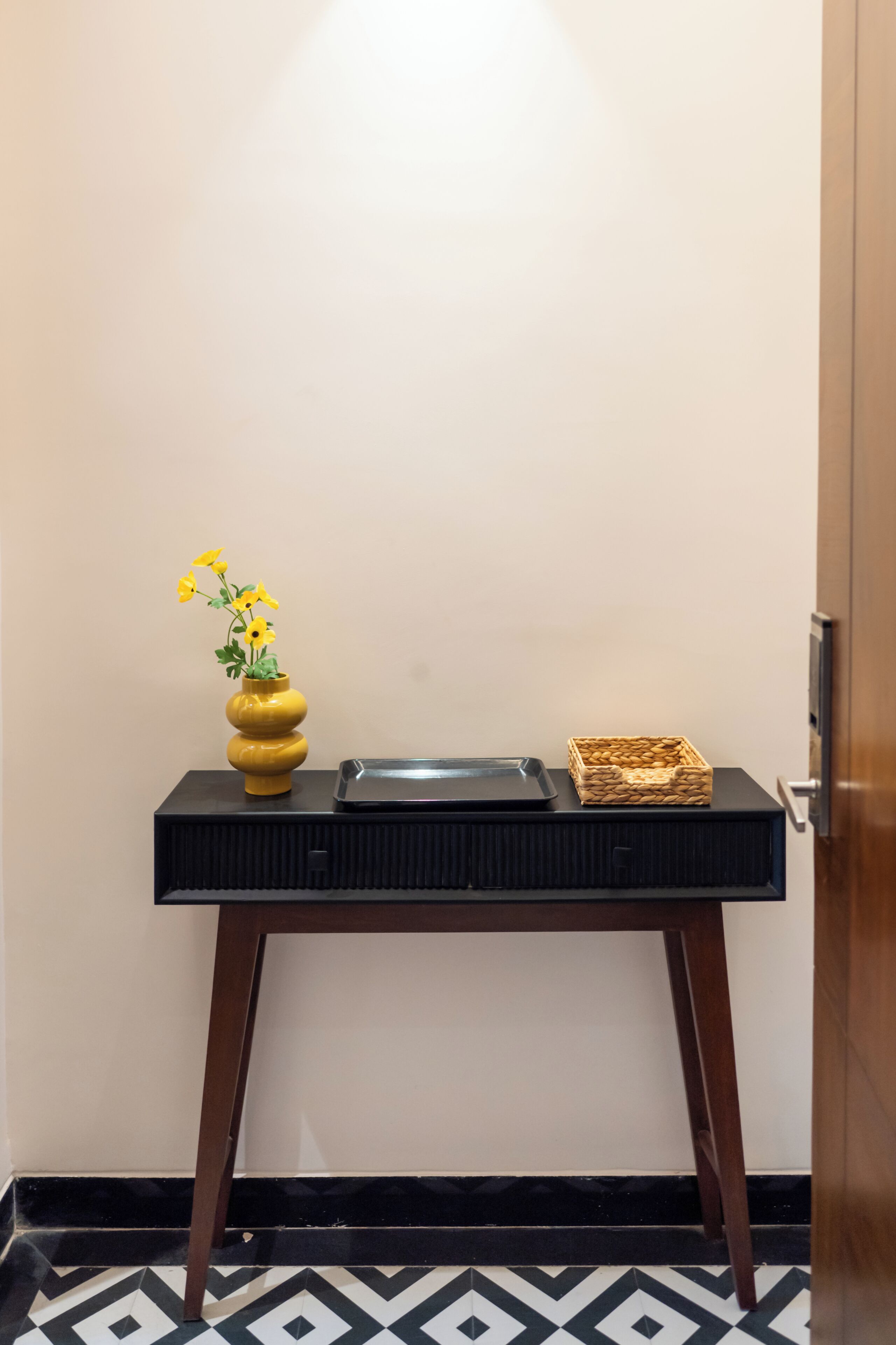 Um aparador preto com um vaso de flores amarelas, um tabuleiro e uma cesta de vime em frente a uma parede lisa.