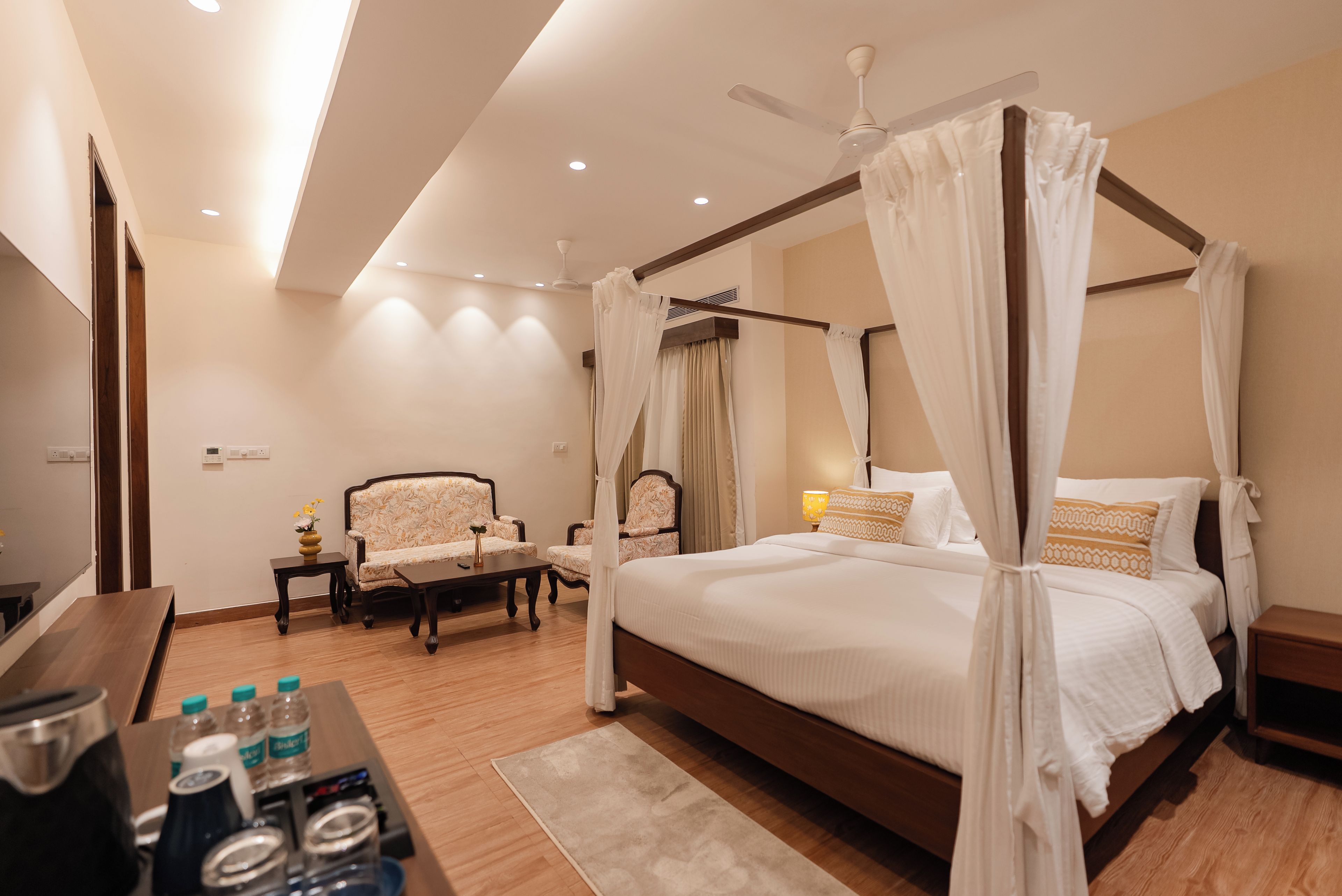 A four-poster canopy bed with a wooden frame and white bedding, a sitting area with a sofa and armchairs, a small table with a vase and bottles, and a ceiling fan.