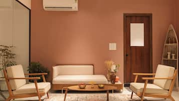 Una sala de estar con un sofá color beige, dos sillas de madera, una mesa de centro, una puerta de madera y una unidad de aire acondicionado en la pared.