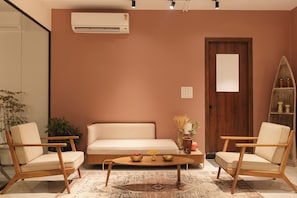 Una sala de estar con un sofá color beige, dos sillas de madera, una mesa de centro, una puerta de madera y una unidad de aire acondicionado en la pared.