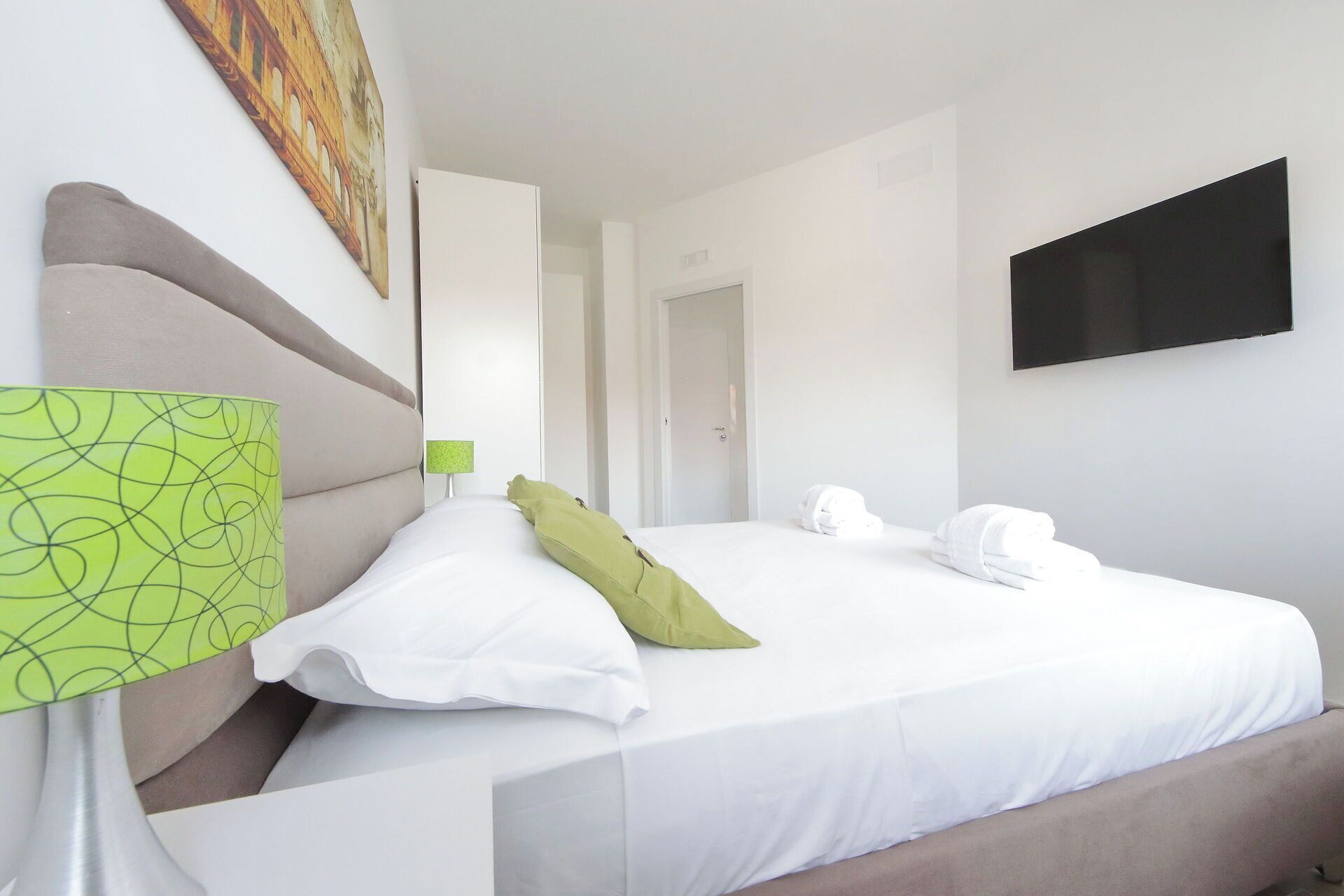 A hotel room with a bed, a TV mounted on the wall, and a painting above the bed.