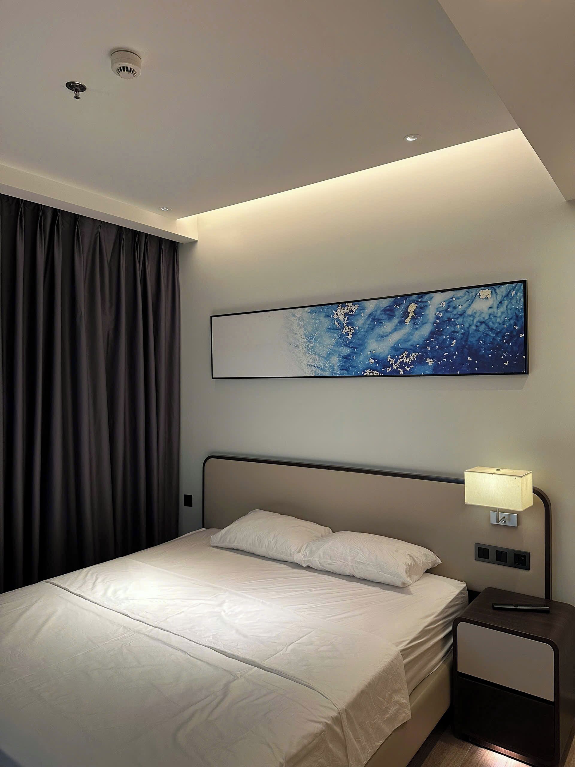 A modern hotel room with a bed, a nightstand, a wall-mounted lamp, a ceiling light, and a large abstract painting.