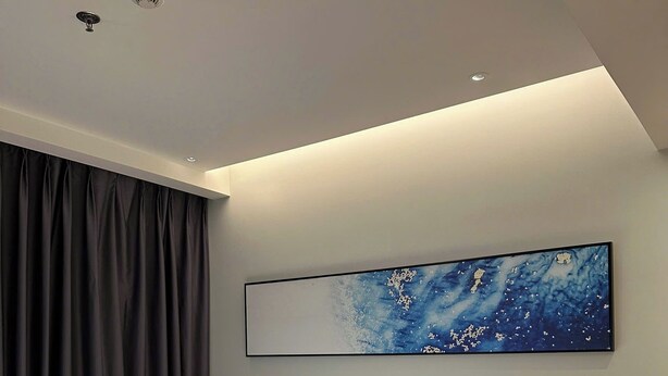 Une chambre d’hôtel moderne, équipée d’un lit, d’une table de chevet, d’une lampe fixée au mur, d’un luminaire au plafond et d’un grand tableau abstrait.
