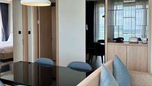 A modern hotel room with a large dining table, blue upholstered chairs, a sofa with blue cushions, a hanging light fixture, and a view of the city through a window. - CMH Homestay A side - 5star standard (Halong City)