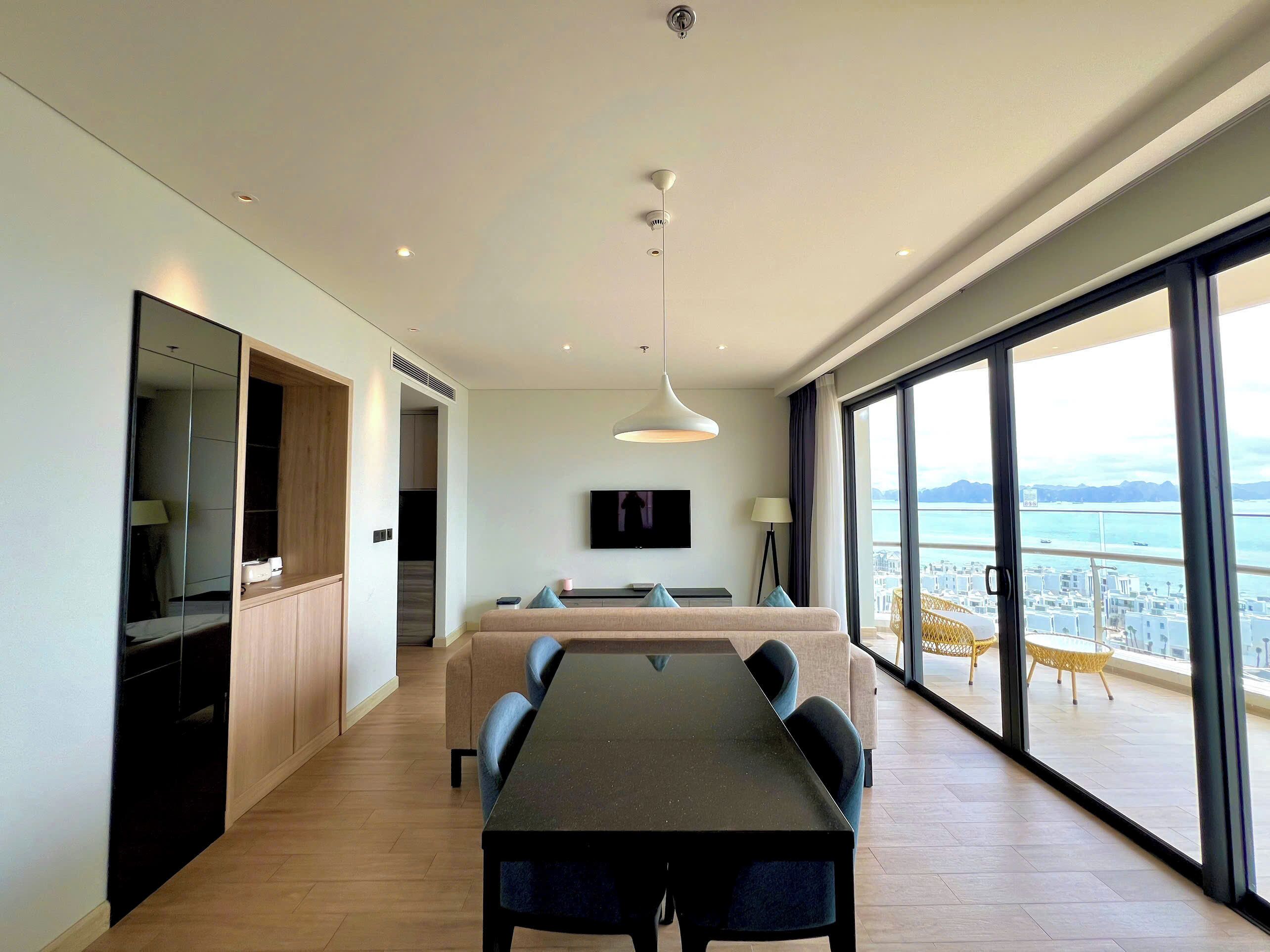 A modern hotel room with a dining area, a large window with a city view, and a balcony with outdoor furniture.