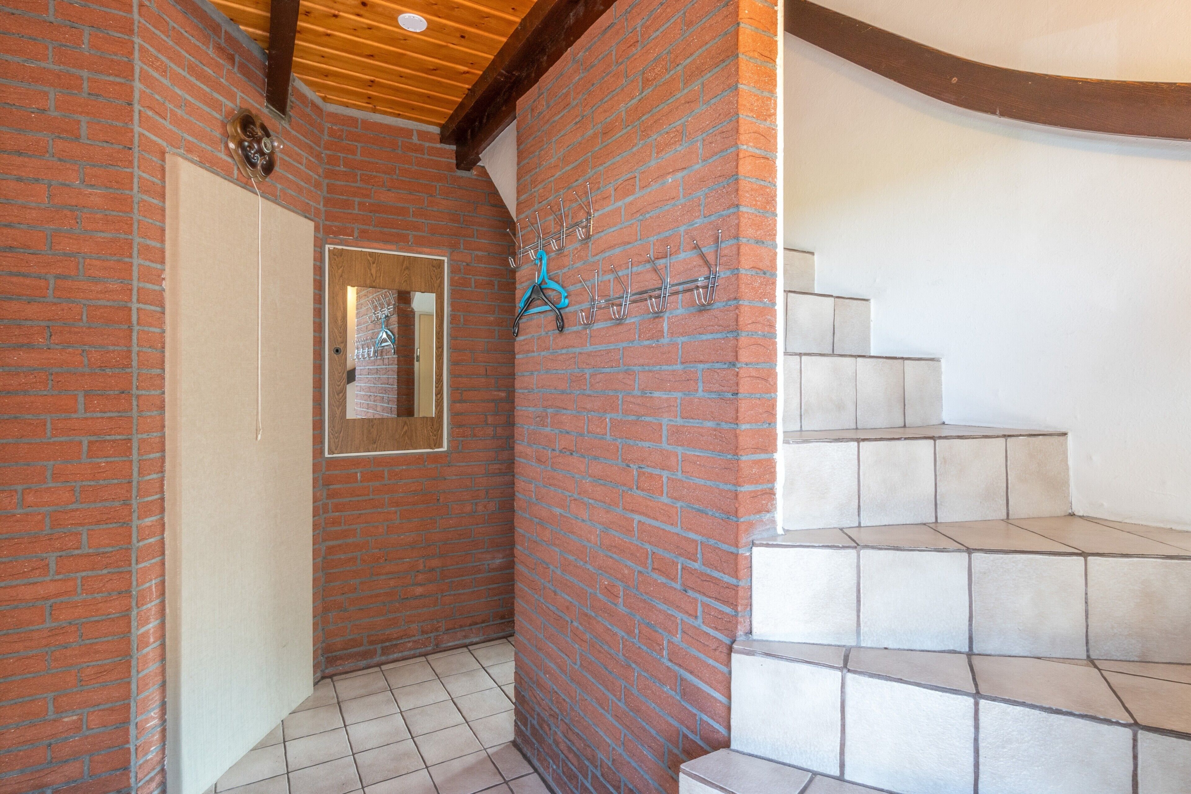 Een hal met houten plafond, bakstenen muur, een deur en een betegelde trap.