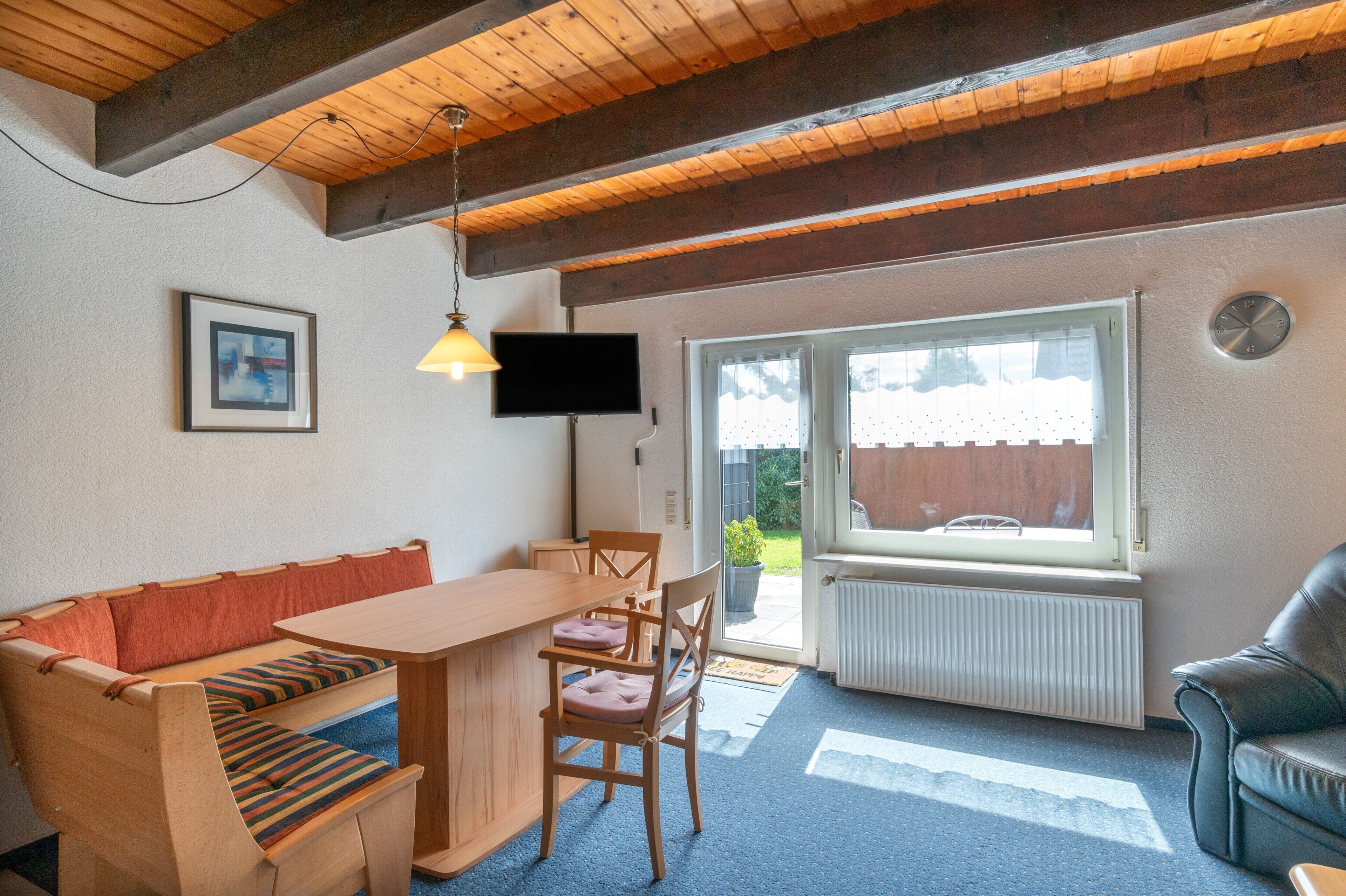 Ein gemütliches Zimmer mit Holzbalkendecke, einer Couch, einem Esstisch, Stühlen, einem Fernseher und einem Fenster mit Blick ins Freie.
