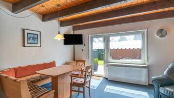 Ein gemütliches Zimmer mit Holzbalkendecke, einer Couch, einem Esstisch, Stühlen, einem Fernseher und einem Fenster mit Blick ins Freie.
