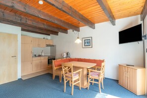 A kitchen with wooden cabinets, a dining table with chairs, a bench, a television, and a ceiling with exposed beams.