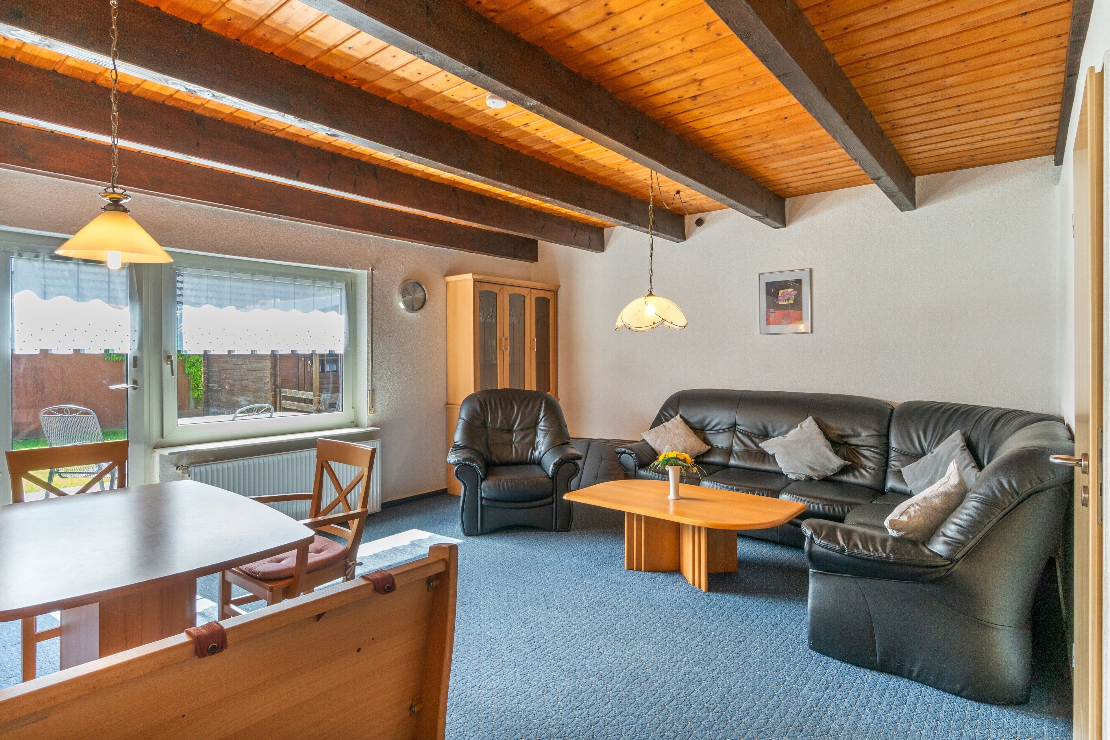 Een gezellige woonkamer met een houten plafond, een zwarte leren bank, een houten salontafel, en een eethoek met tafel en stoelen.