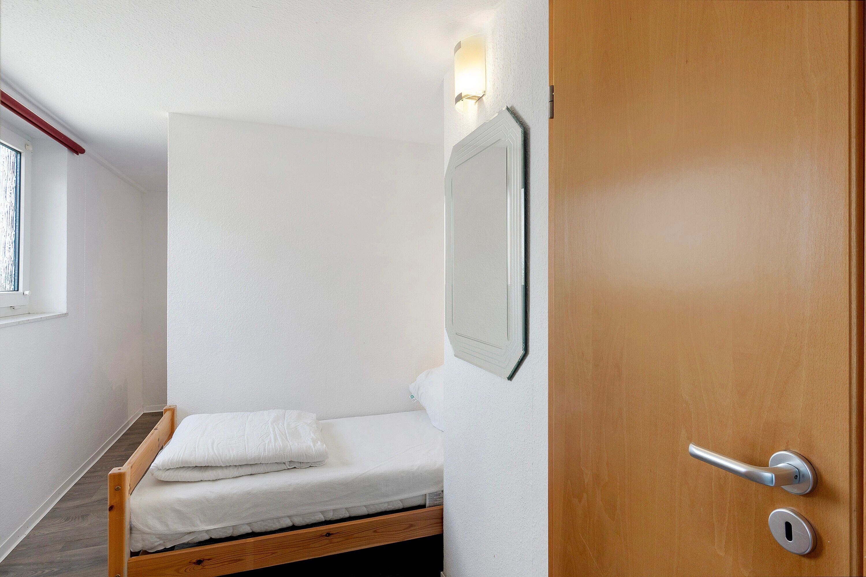 Una habitación pequeña con cama individual, piso de madera, pared blanca y puerta con picaporte plateado.