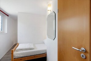 A small, single-bed room with a wooden floor, a white wall, and a door with a silver handle.