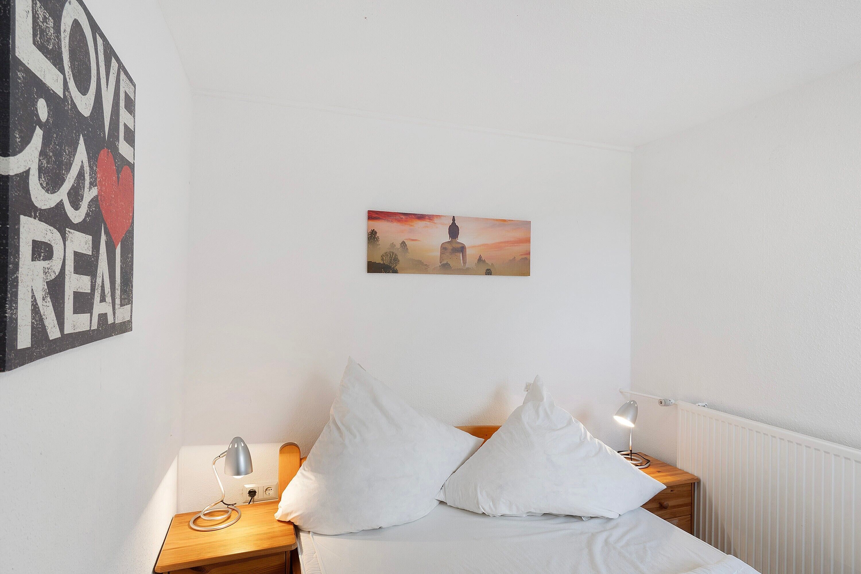 Ein Schlafzimmer mit einem Bett, zwei Nachttischlampen, einem Wandbild und einem Poster mit Text und Herzsymbol.