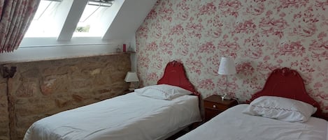 A room with two beds, a stone wall, a window with curtains, and a floral wallpaper.
