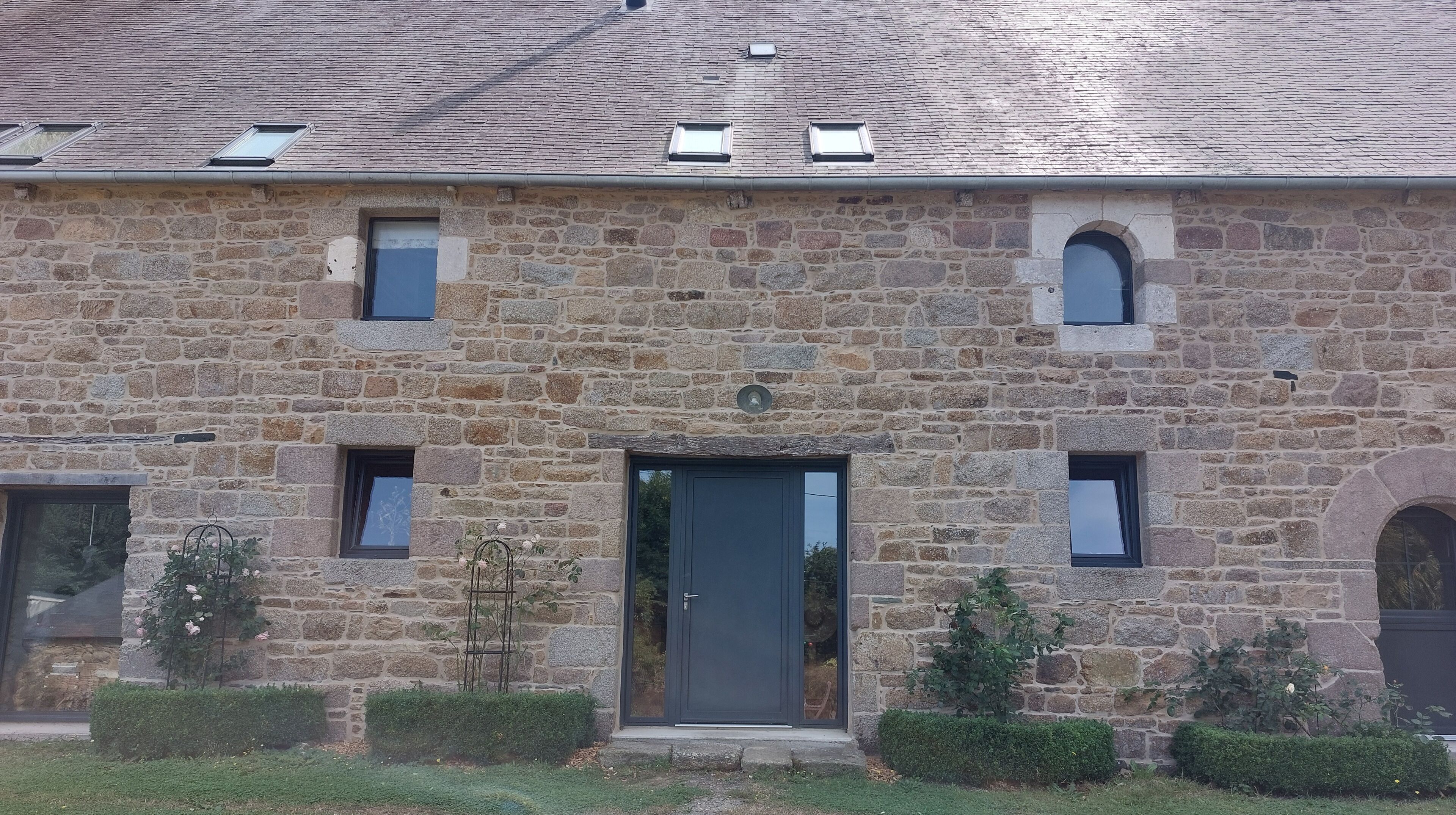 A stone building with a dark entrance door, multiple windows, and a small garden area with plants.
