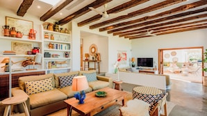 A cozy living room with a sofa, coffee table, and a TV. There are shelves with decorative items and a wooden ceiling.