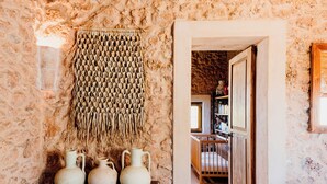 A rustic interior with a woven wall hanging, a wooden shelf with pottery, and a doorway leading to a room with a bookshelf.