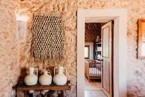 A rustic interior with a woven wall hanging, a wooden shelf with pottery, and a doorway leading to a room with a bookshelf.