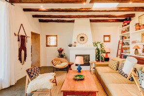 A cozy living room with a fireplace, wooden beams, a woven chair, a sofa, and a bookshelf.