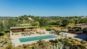 A pool area with lounge chairs and umbrellas, surrounded by greenery and a view of the landscape.