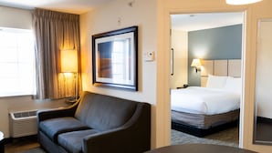 Une chambre d’hôtel avec un canapé, un lit, un tableau encadré au mur, une lampe et une fenêtre avec des rideaux.