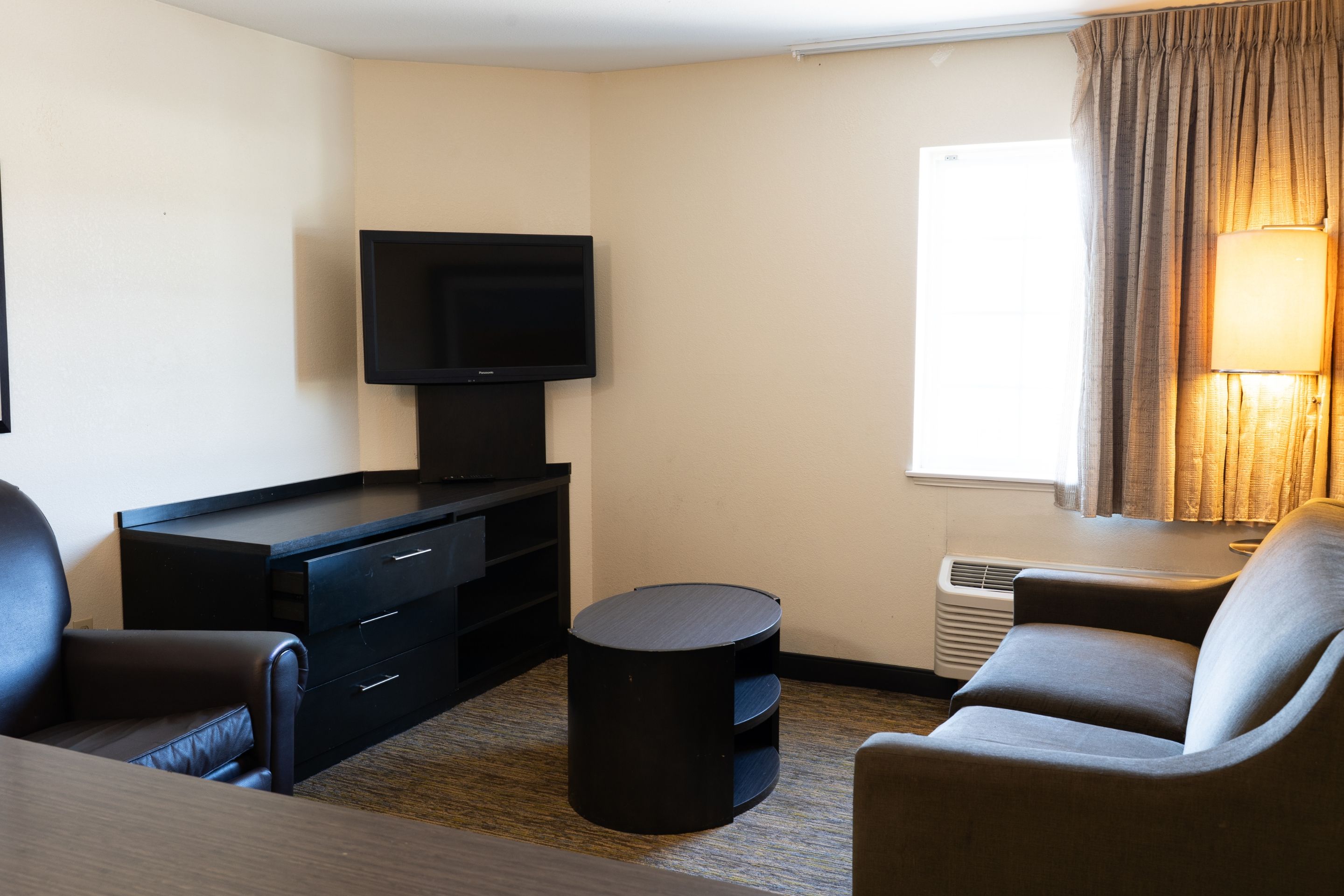 Habitación de hotel con televisión de pantalla plana, un escritorio con cajones, una mesa redonda, dos sillones, una ventana con cortinas y un calentador de pared.