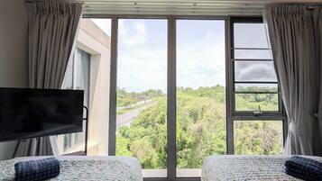 Habitación de hotel con dos camas, televisor y ventana grande que ofrece vistas a un paisaje verde.