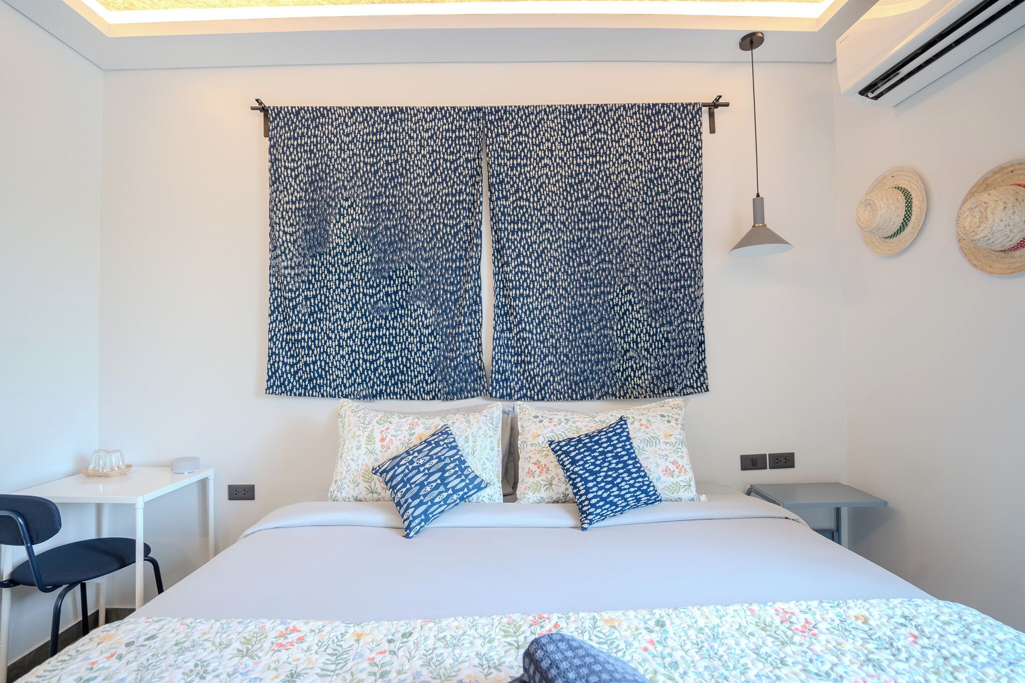 Quarto com cama, duas mesas de cabeceira, escrivaninha, cadeira e janela com cortinas estampadas.