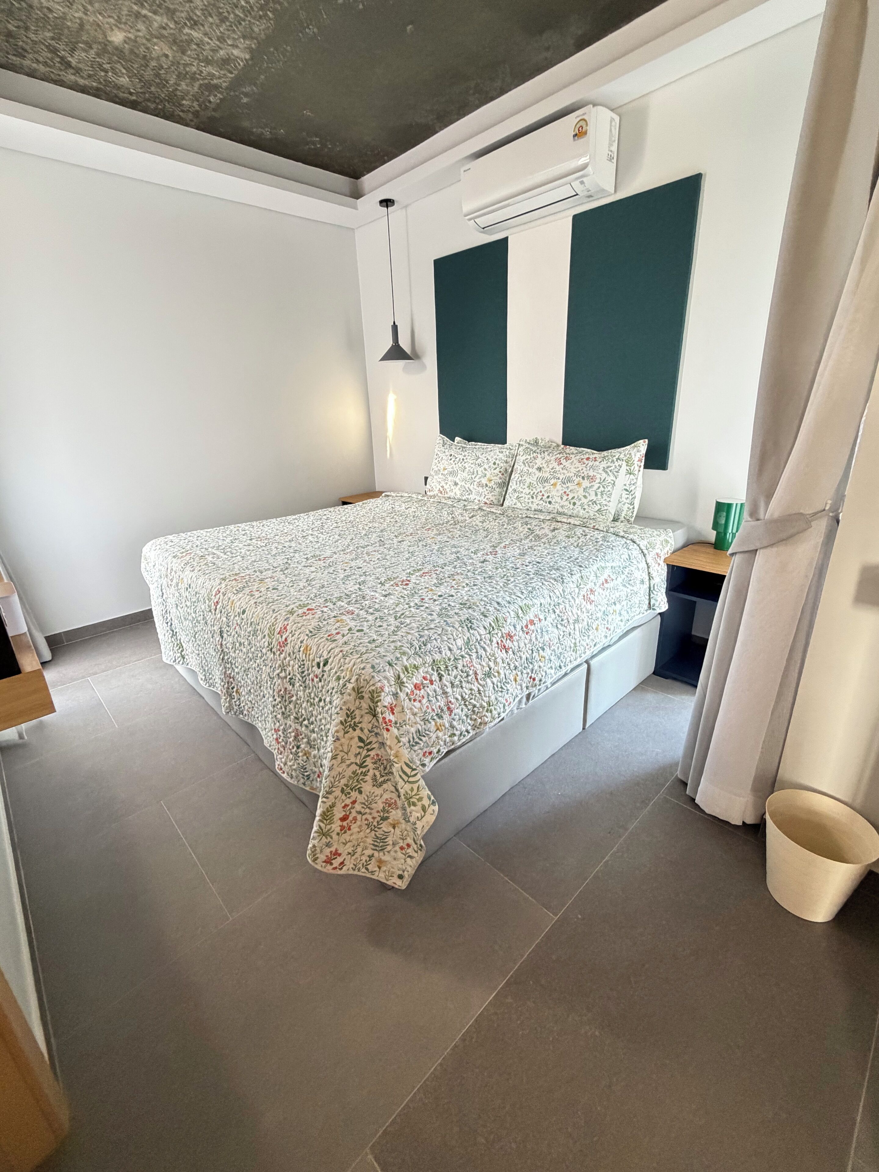A bedroom with a bed, an air conditioning unit, a bedside table, and a curtain.