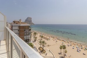 Ein Strand mit Palmen und Sonnenschirmen, ein Felsvorsprung in der Ferne und ein Gebäude mit Balkonen mit Blick auf den Strand.