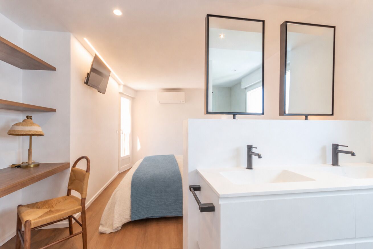 A modern bathroom with a double sink, two mirrors, a chair, a bedside table, and a television mounted on the wall.