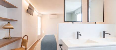 A modern bathroom with a double sink, two mirrors, a chair, a bedside table, and a television mounted on the wall.