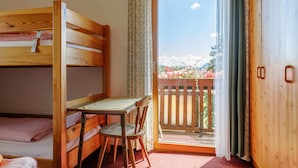 A room with a bunk bed, a desk with a chair, and a balcony with a view of flowers and mountains.