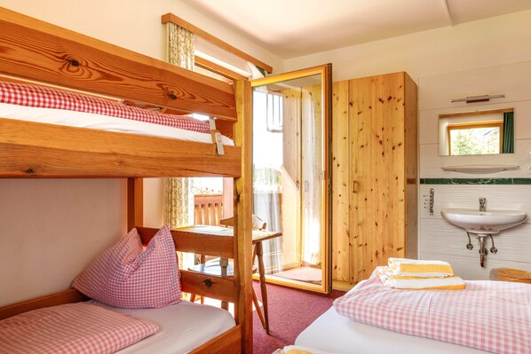 A bunk bed room with a wooden desk and chair, a small balcony, and a bathroom with a sink.