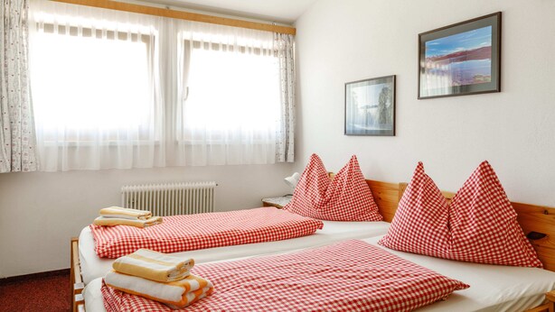 A room with two beds, each with red and white checkered bedding and pillows, a radiator, and two framed pictures on the wall.