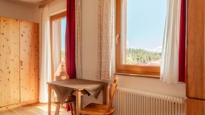 A room with a wooden floor, a table with a tablecloth, two chairs, a window with a view of trees, and a radiator.