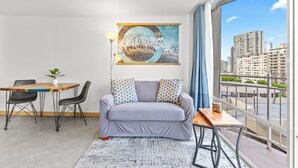 A modern living room with a sofa, a dining table, and a view of the cityscape through large windows. - Ilikai Marina - Modern Designer Studio (Honolulu)