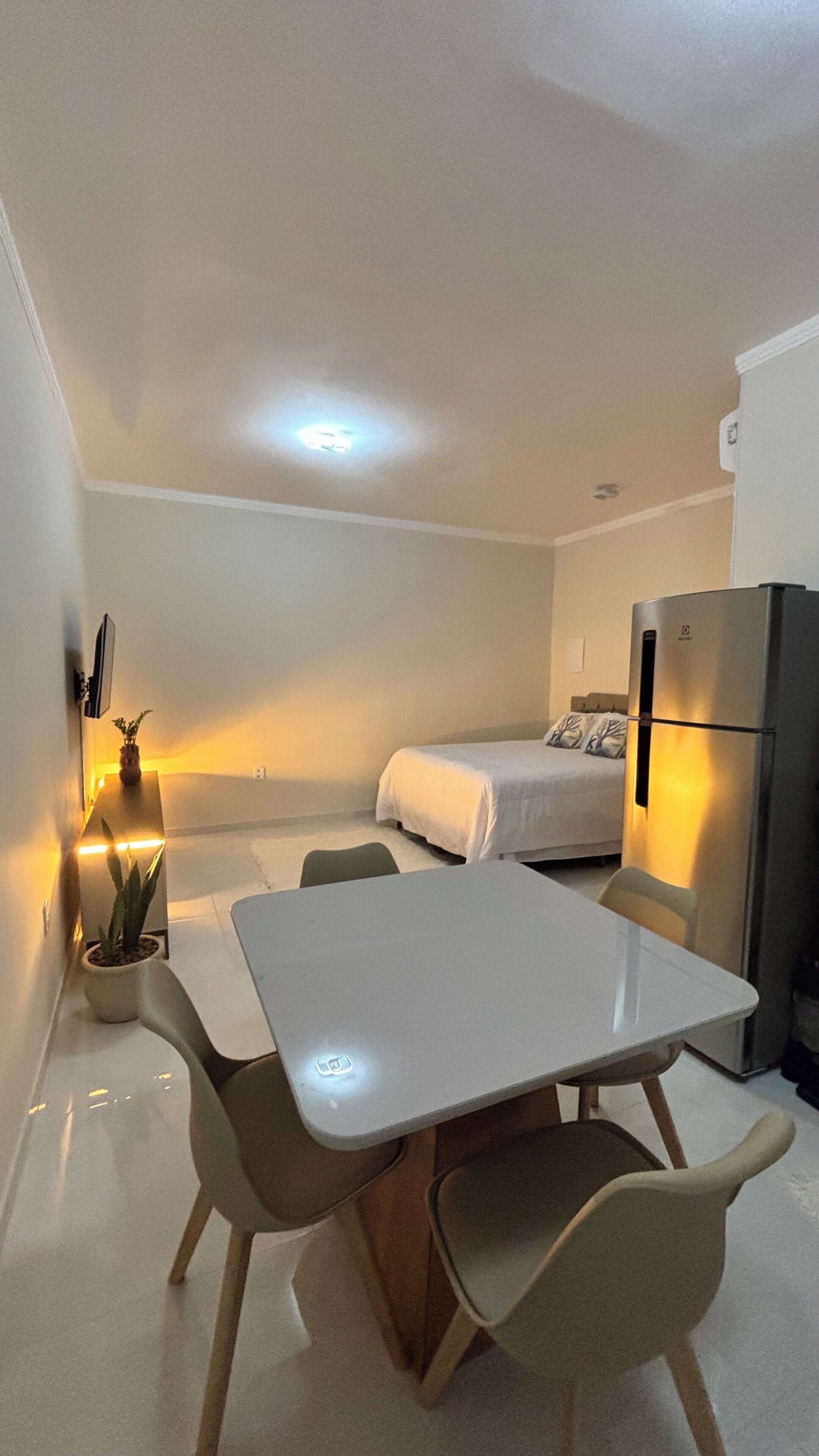 A compact room with a bed, a dining table with chairs, a refrigerator, and a potted plant.