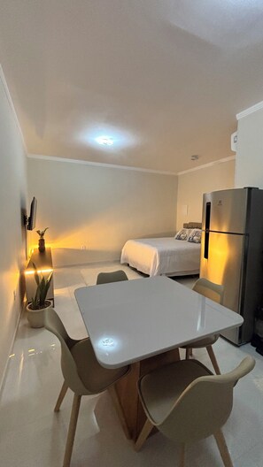 Quarto compacto com cama, mesa de jantar com cadeiras, geladeira e um vaso de planta.