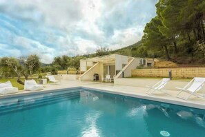 Una piscina con sillas de descanso, un pequeño edificio de techo inclinado y un paisaje con árboles al fondo.