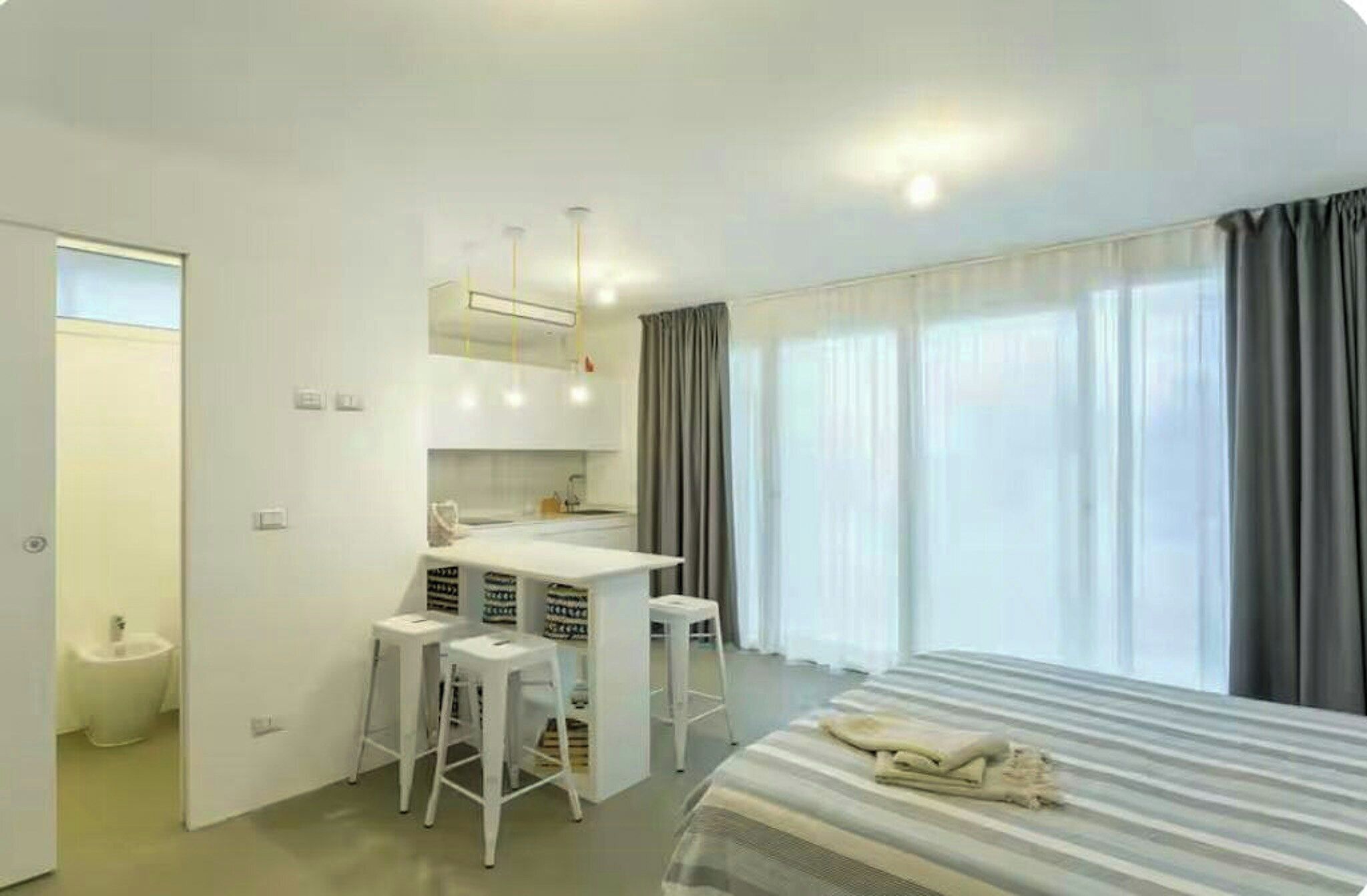 Habitación de hotel con cama, escritorio con silla, un taburete pequeño, espejo y ventana con cortinas.