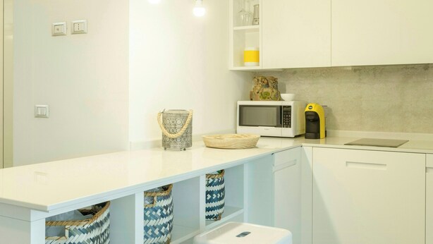 Uma cozinha moderna com armários brancos, uma ilha central com banquetas, iluminação pendente e um micro-ondas na bancada.