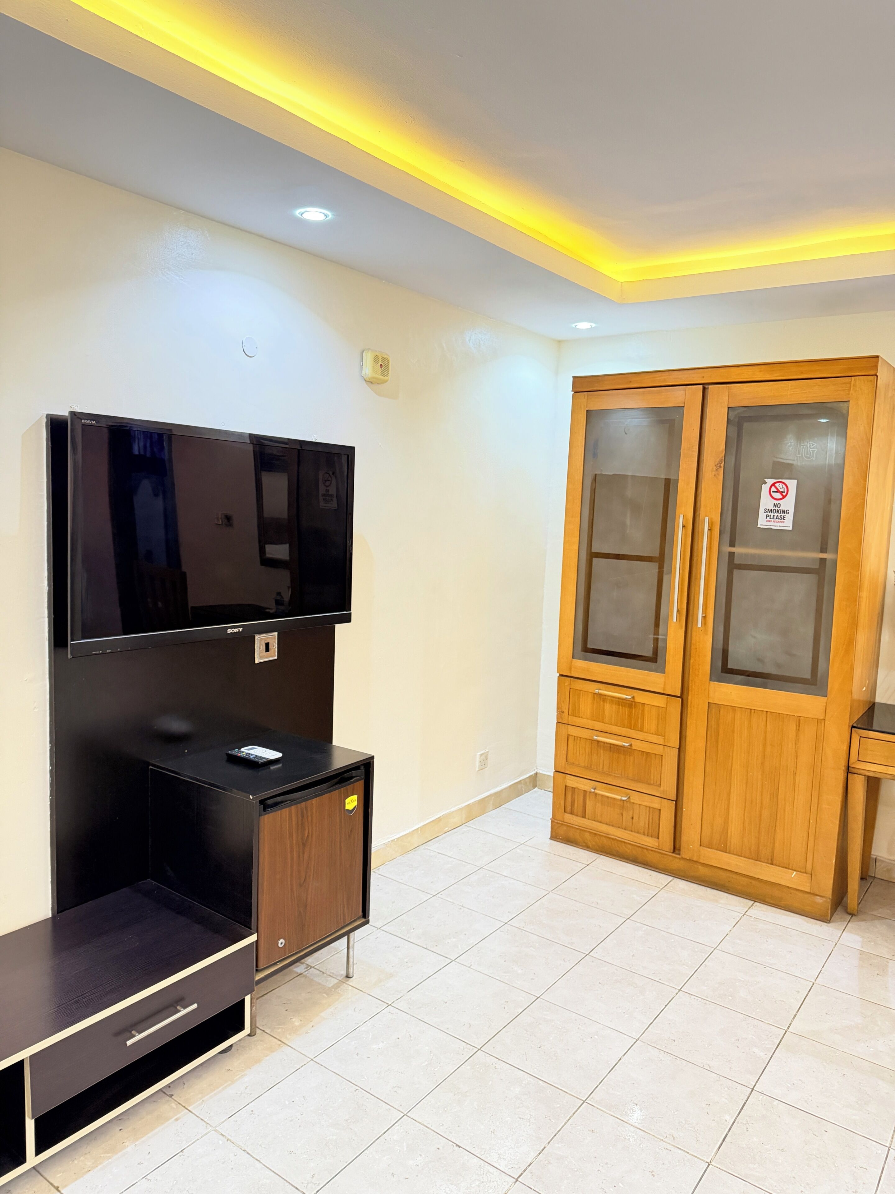 A room with a flat-screen TV mounted on a black entertainment unit, a wooden wardrobe with glass doors, and a 'No Smoking' sign on the wardrobe.