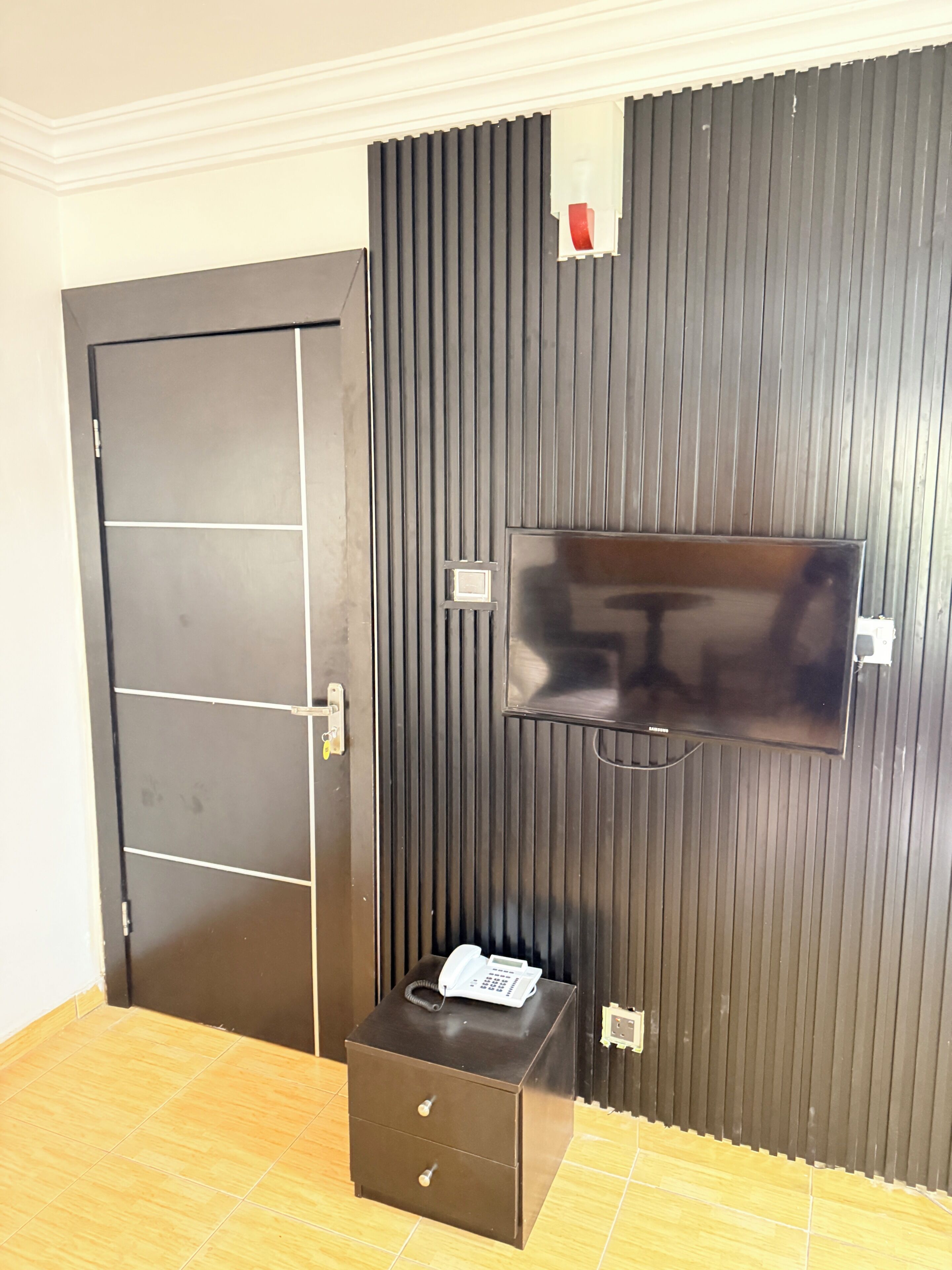 A room with a closed door, a flat-screen TV mounted on a dark wall, and a small wooden chest of drawers.